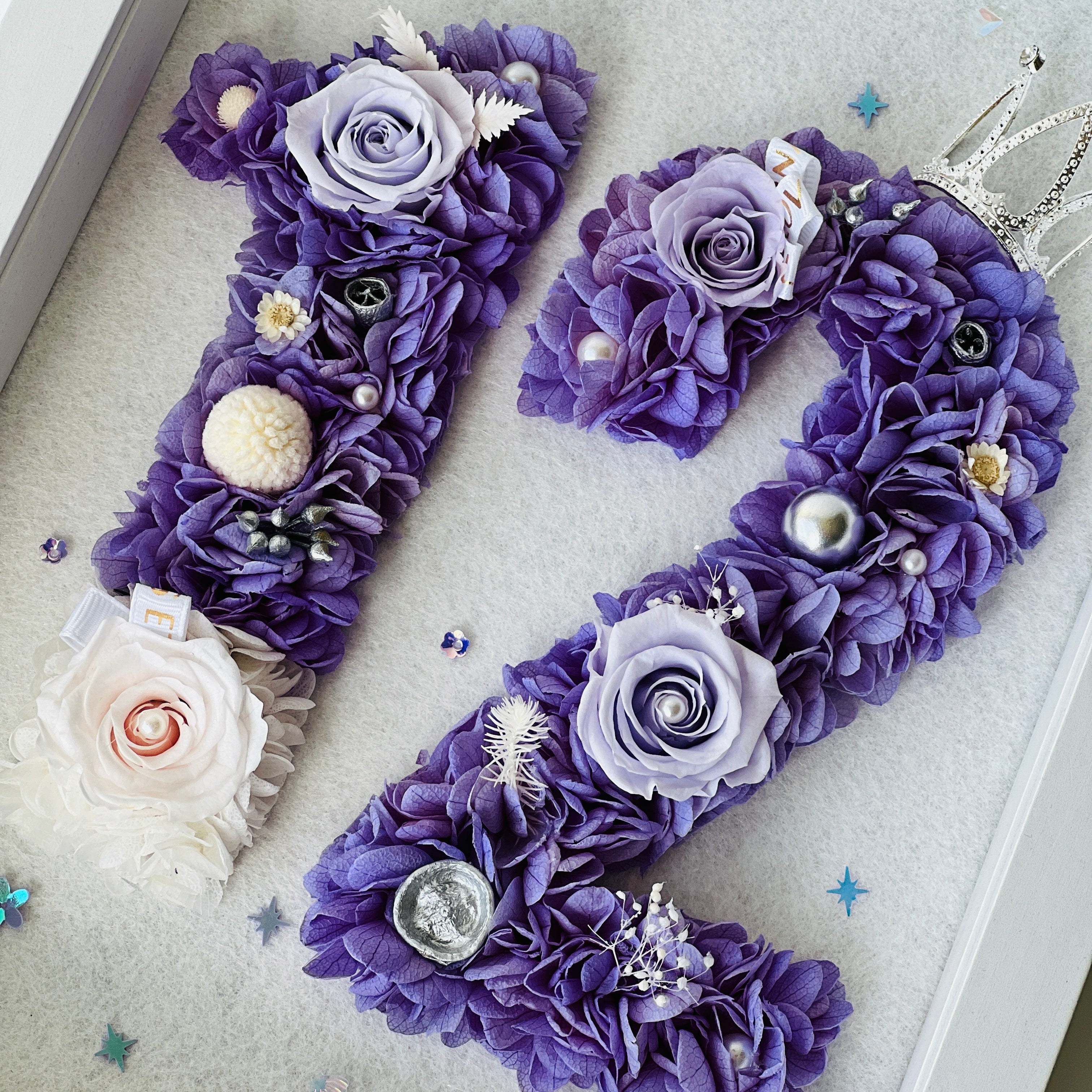 Custom number floral artwork featuring preserved roses and hydrangeas in regal purple and white, accented with sparkling embellishments and a tiara. Perfect for commemorating milestones.