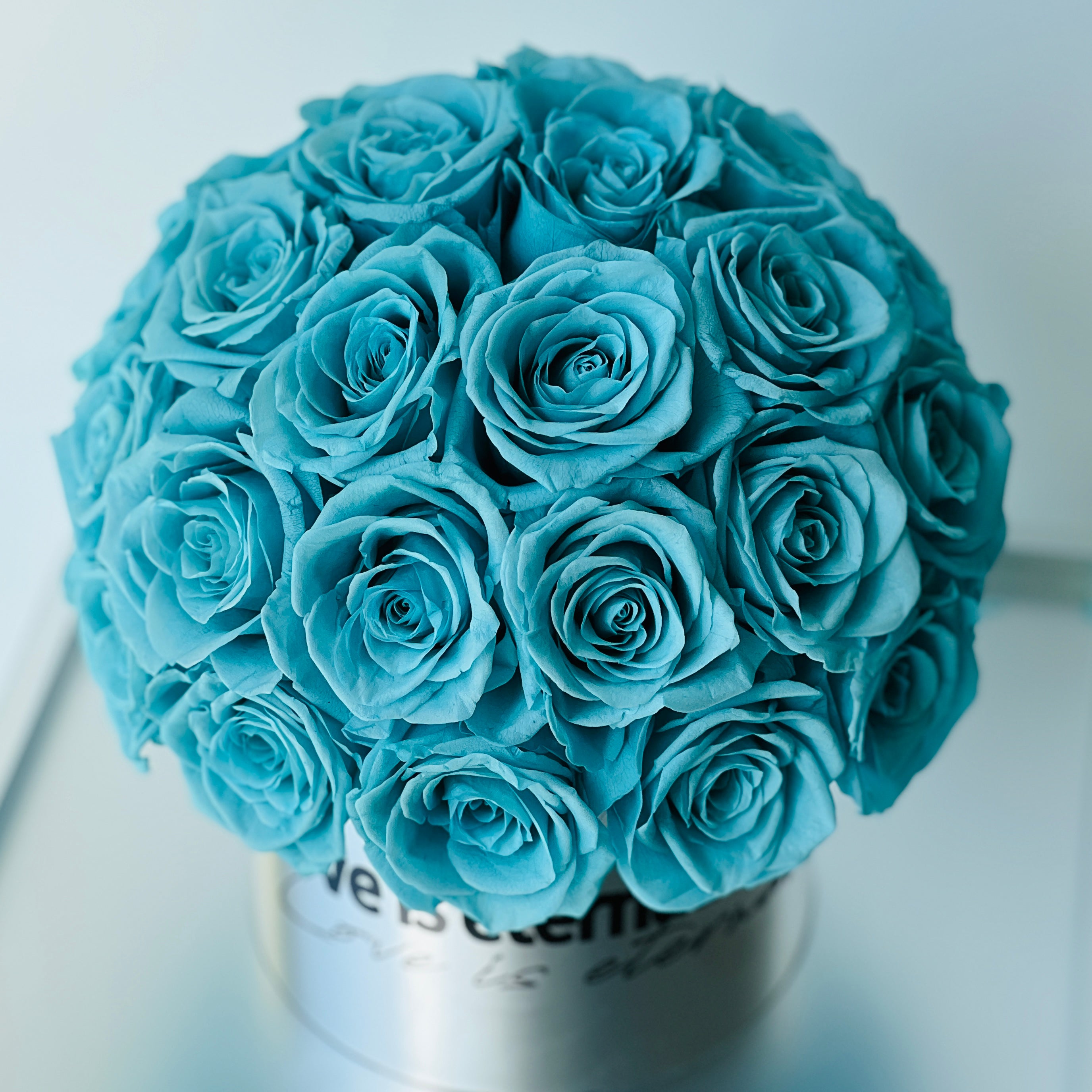 The Tiffany Blue Rose Hatbox features preserved roses on a sleek grey base inscribed with "love is eternal." This serene eternity rose arrangement offers a modern statement of enduring affection, perfect for adding tranquility to any space or as a heartfelt gift.