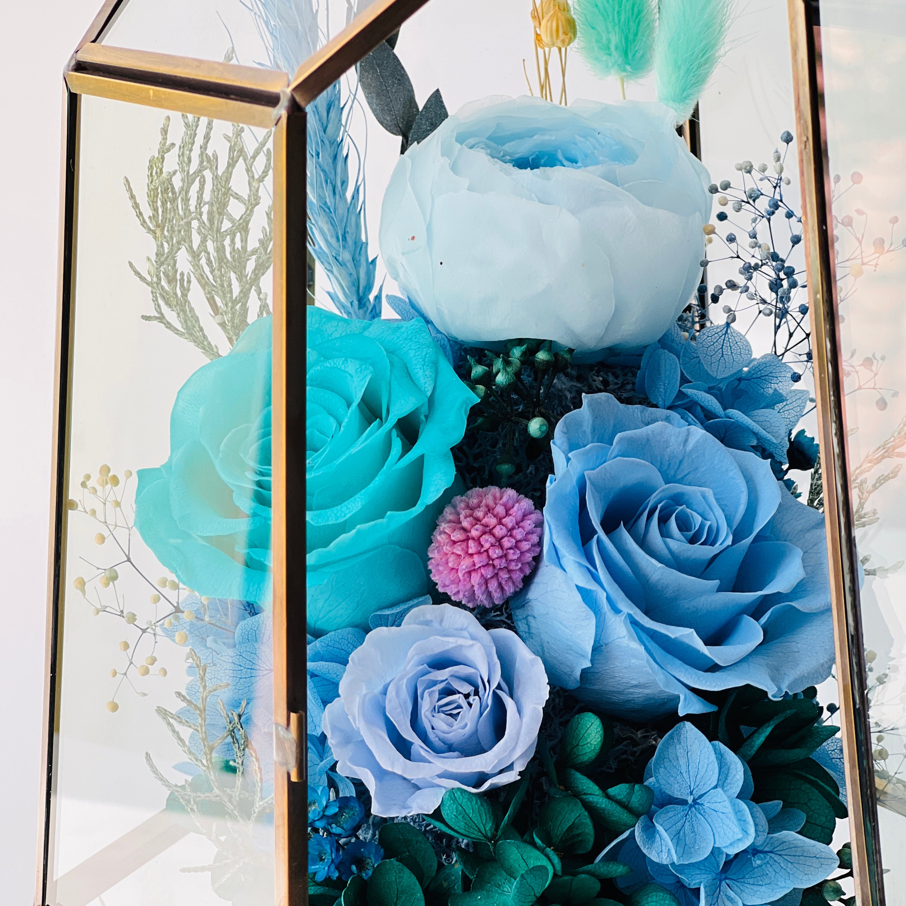 Exquisite Ocean Breeze preserved floral terrarium featuring deep ocean blue roses and hydrangeas, accented with greenery and pink florals in a modern geometric vessel. Perfect as a timeless eternity rose gift.