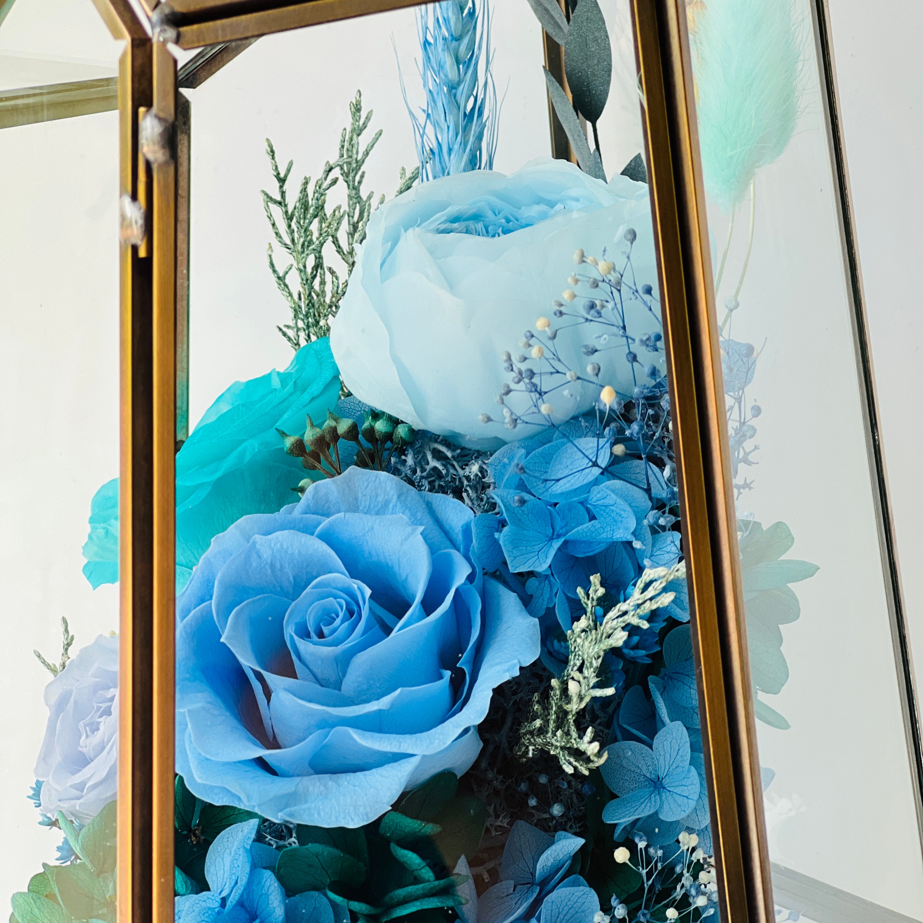 Exquisite Ocean Breeze preserved floral terrarium featuring deep ocean blue roses and hydrangeas, accented with greenery and pink florals in a modern geometric vessel. Perfect as a timeless eternity rose gift.