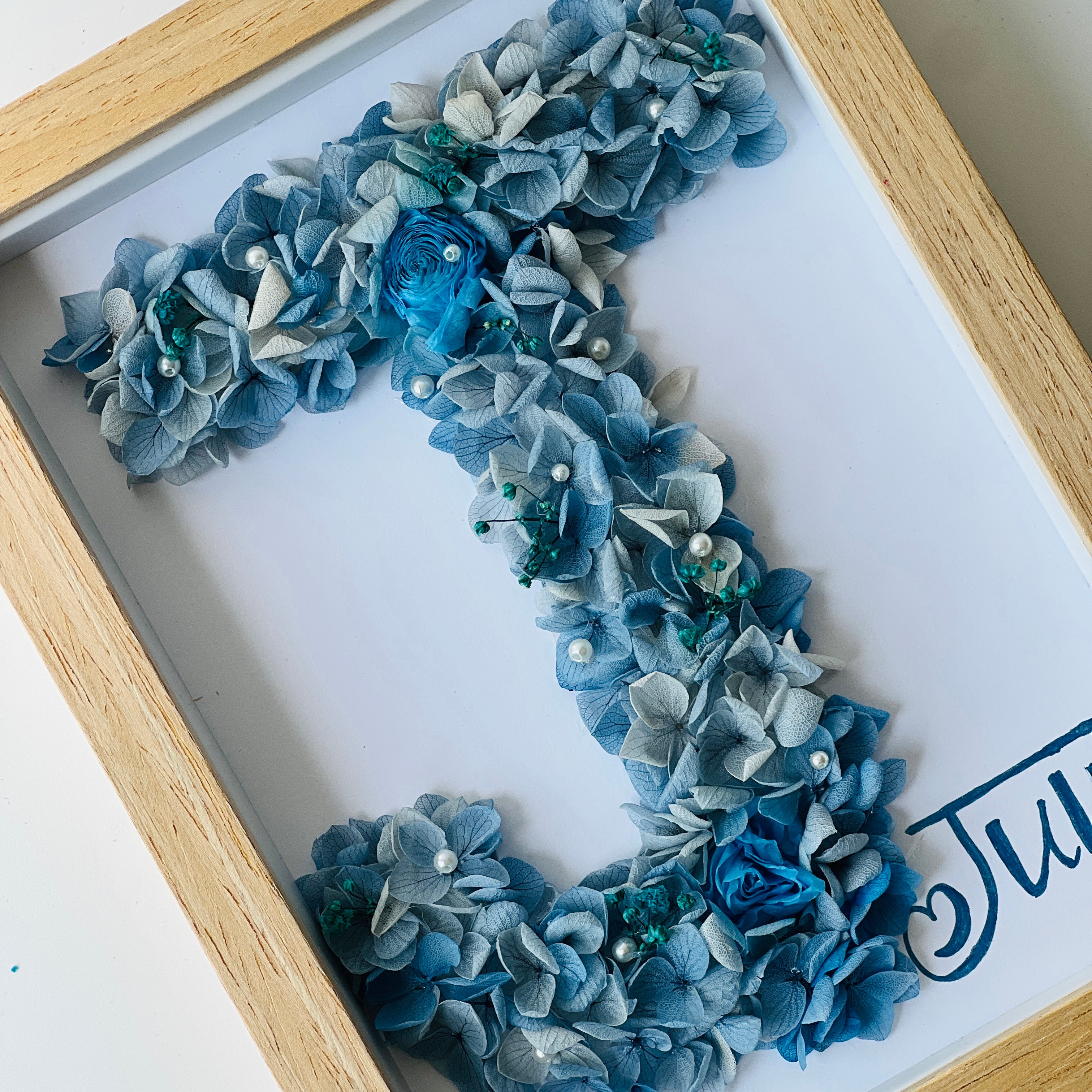 Crafted initial-shaped preserved floral letter art featuring blue and white hydrangeas, with a white rose and pearl accents, framed in natural wood—a personalized gift.