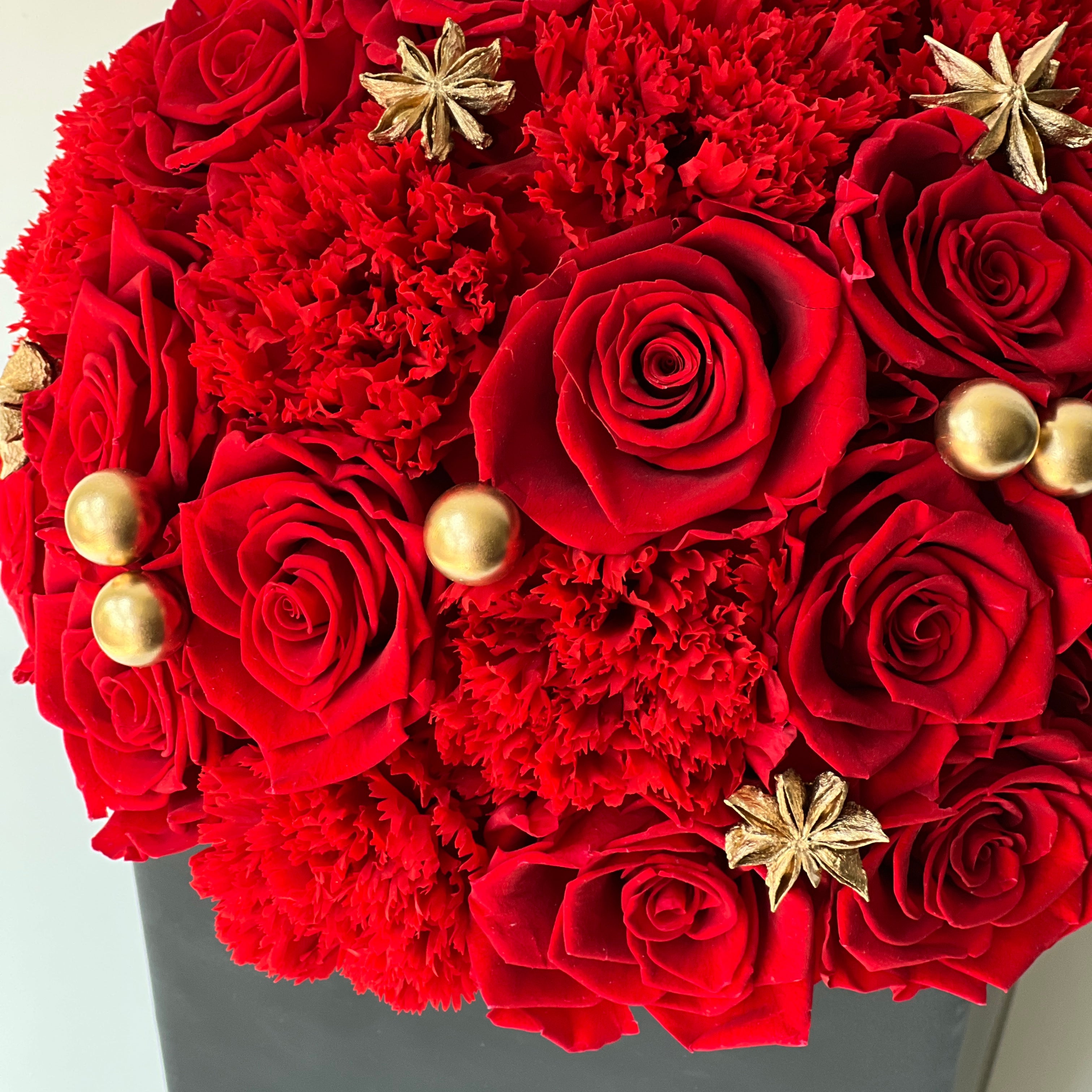 The Regal Red preserved flower arrangement features deep red roses and vibrant carnations, adorned with golden pearls and star-shaped embellishments. Encased in a sleek black box, this luxurious eternity rose gift is perfect for special occasions or as a dramatic centerpiece, celebrating timeless beauty and sophistication year-round.