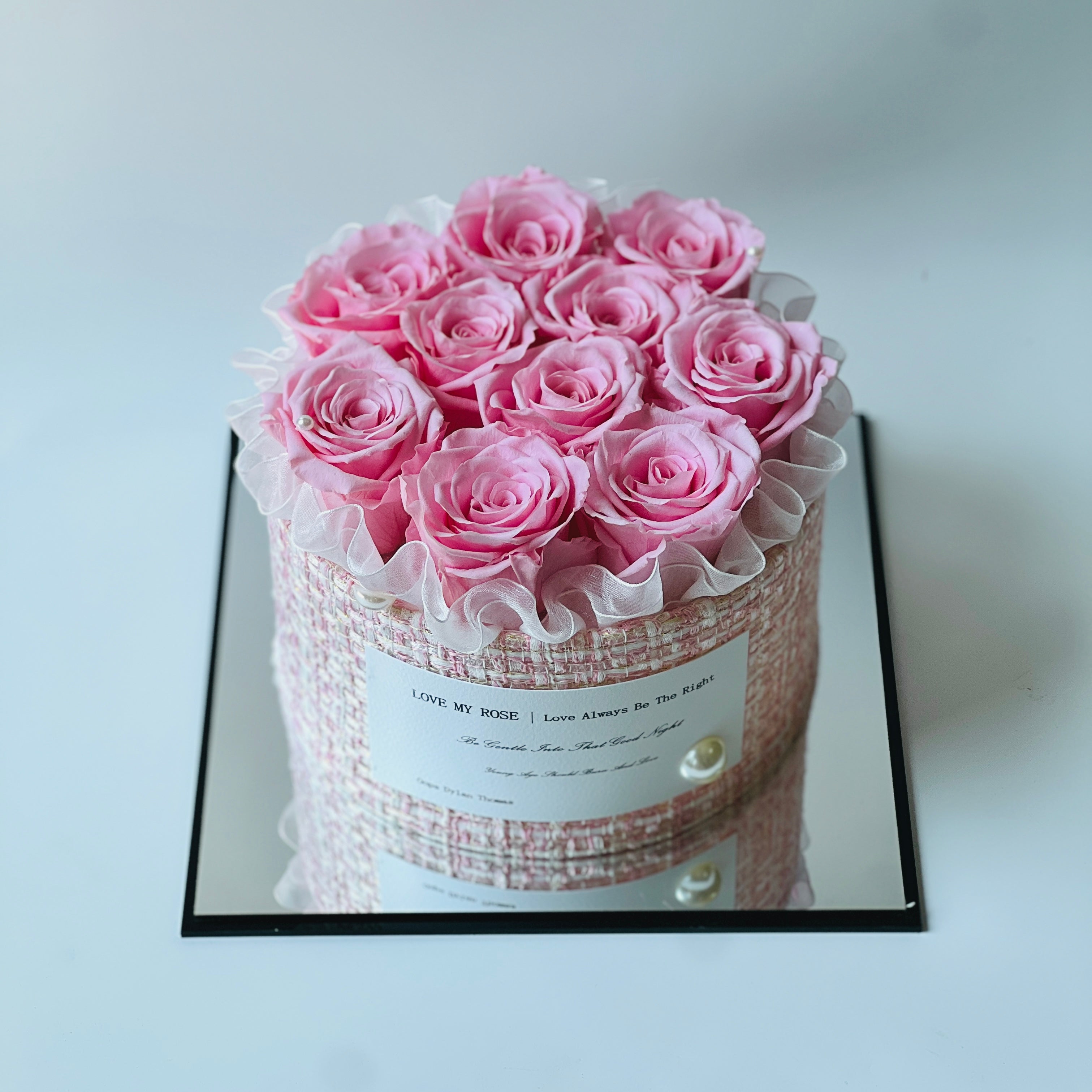 The Pink Perfection preserved flower arrangement features soft pink roses encased in a chic woven pink and white box, adorned with a ruffled ribbon and pearl accents. This timeless eternity rose gift is perfect for adding romance and sophistication to any space.