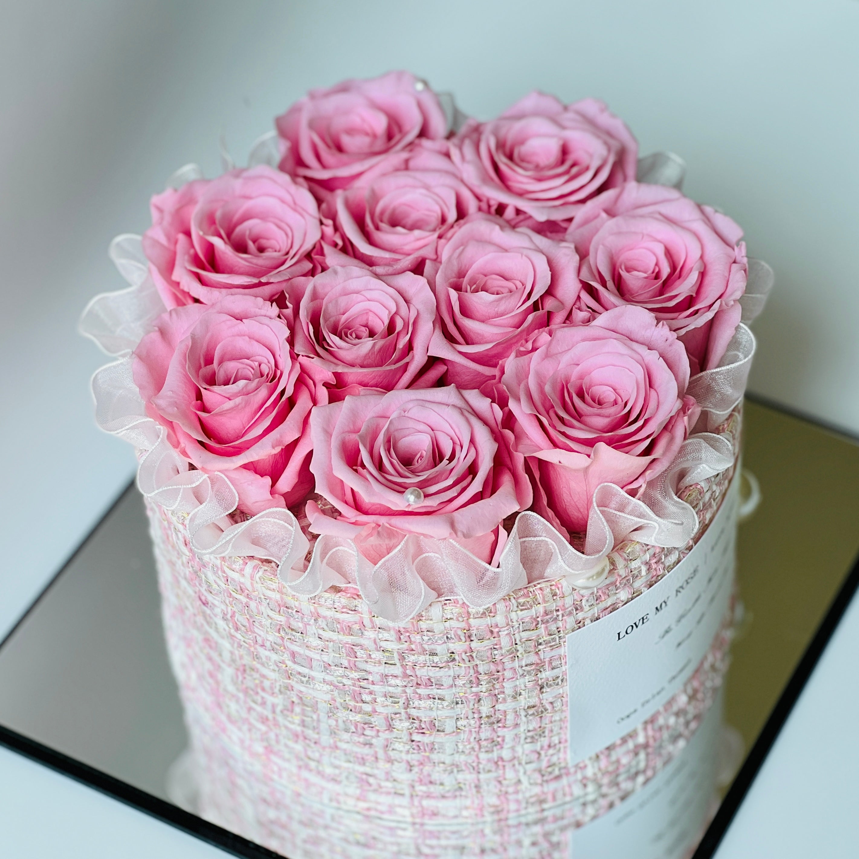 The Pink Perfection preserved flower arrangement features soft pink roses encased in a chic woven pink and white box, adorned with a ruffled ribbon and pearl accents. This timeless eternity rose gift is perfect for adding romance and sophistication to any space.