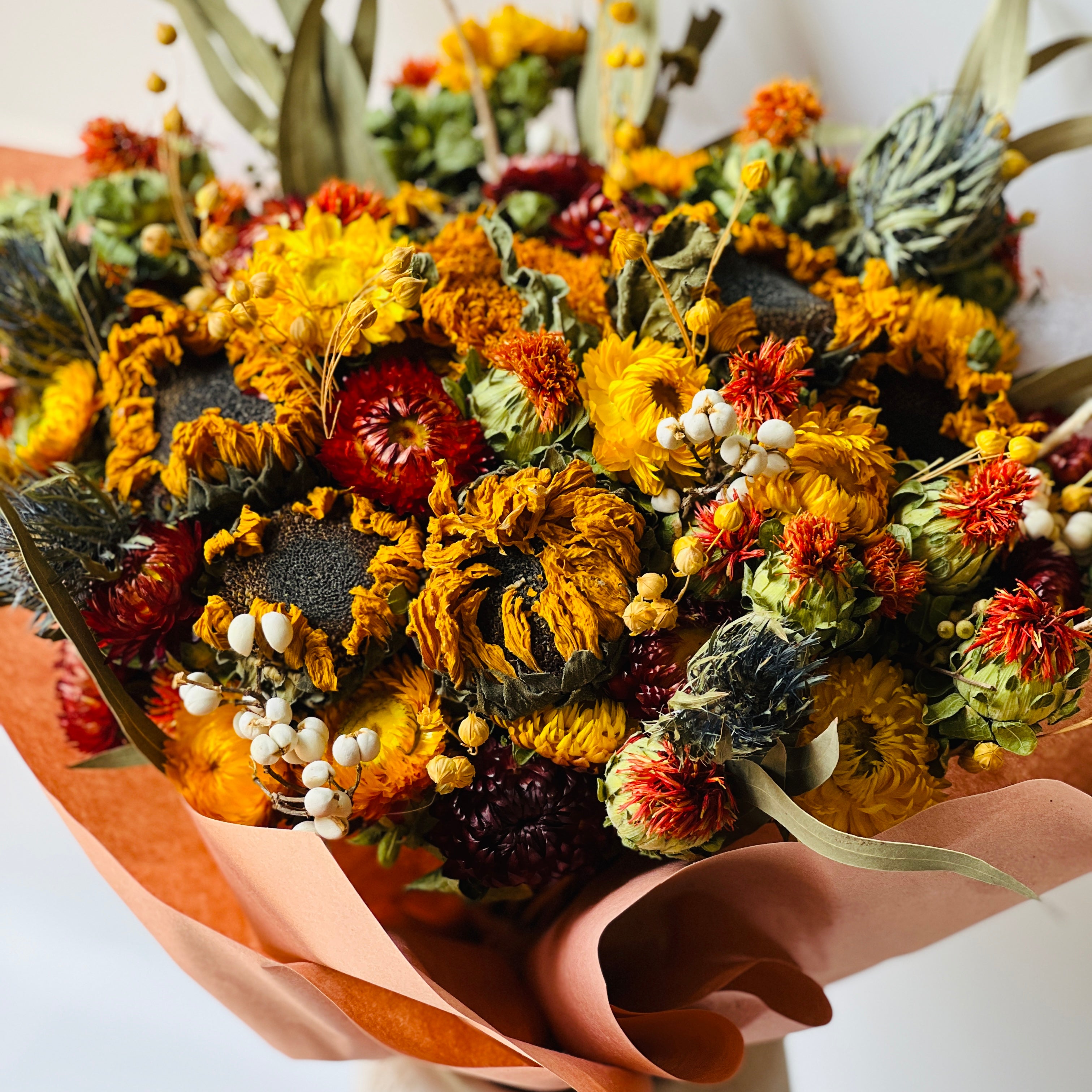 Autumn Harvest Bouquet with sunflowers, craspedia, and strawflowers in warm autumn colors. Accented by soft greenery and wrapped in peach paper, this bouquet adds rustic charm to any space, maintenance-free.