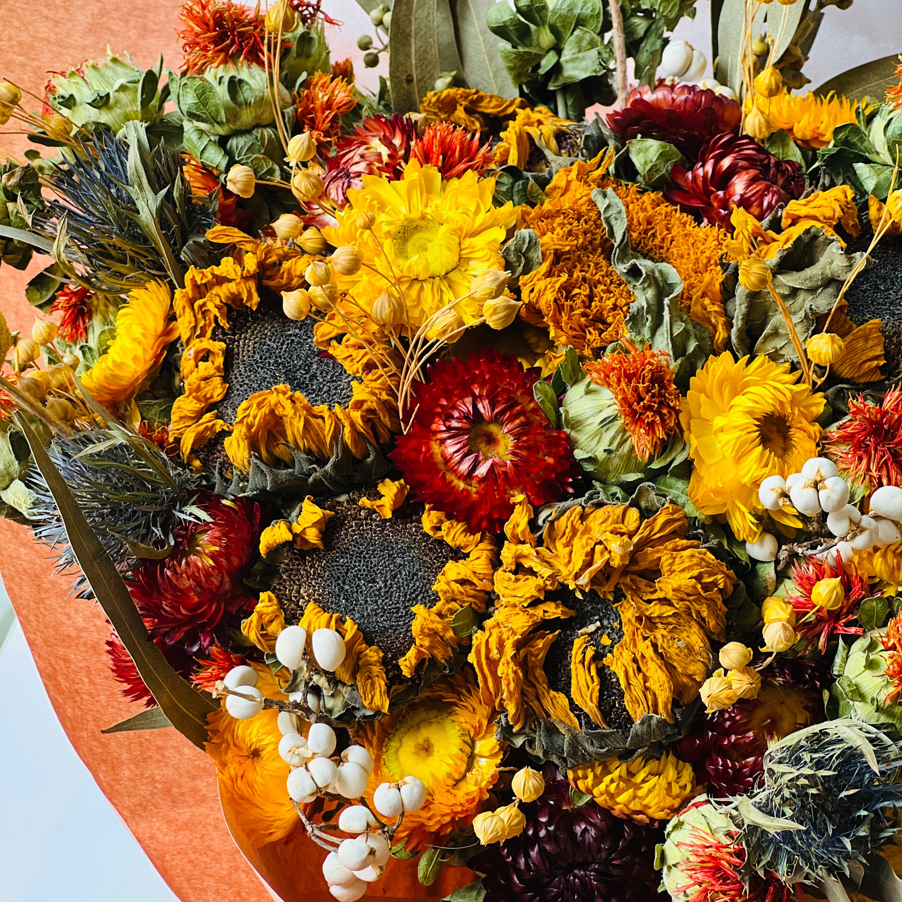 Autumn Harvest Bouquet with sunflowers, craspedia, and strawflowers in warm autumn colors. Accented by soft greenery and wrapped in peach paper, this bouquet adds rustic charm to any space, maintenance-free.