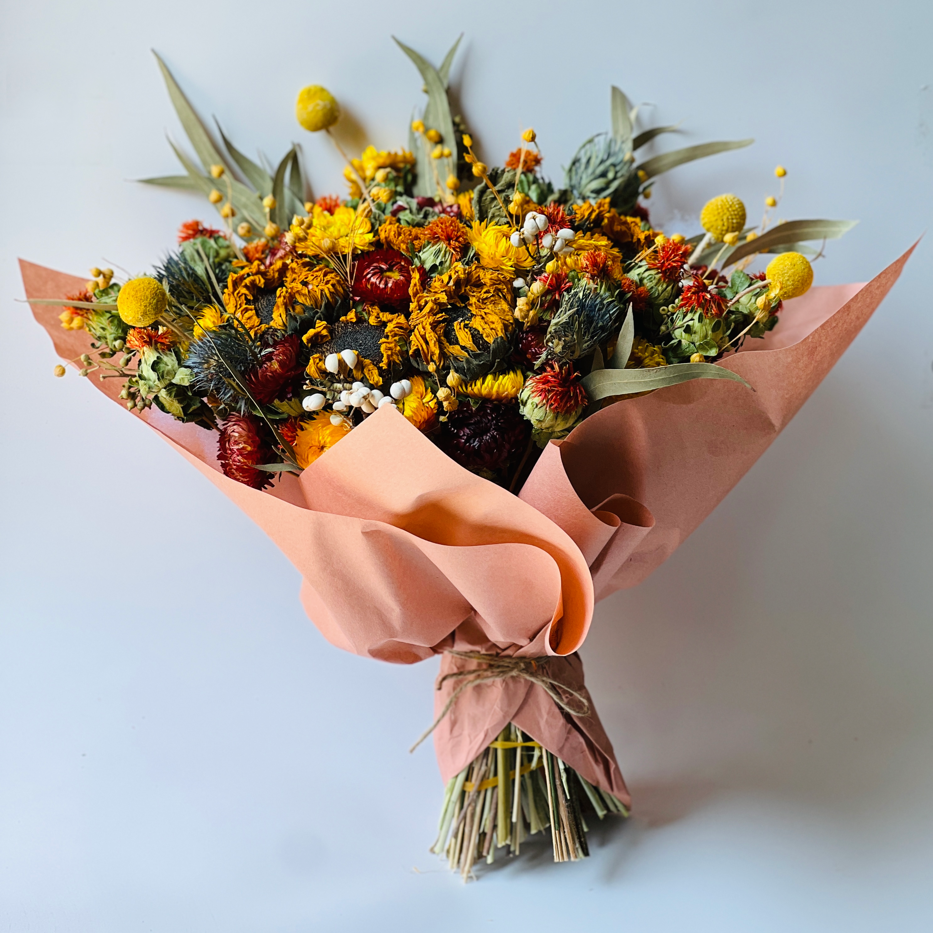 Autumn Harvest Bouquet with sunflowers, craspedia, and strawflowers in warm autumn colors. Accented by soft greenery and wrapped in peach paper, this bouquet adds rustic charm to any space, maintenance-free.