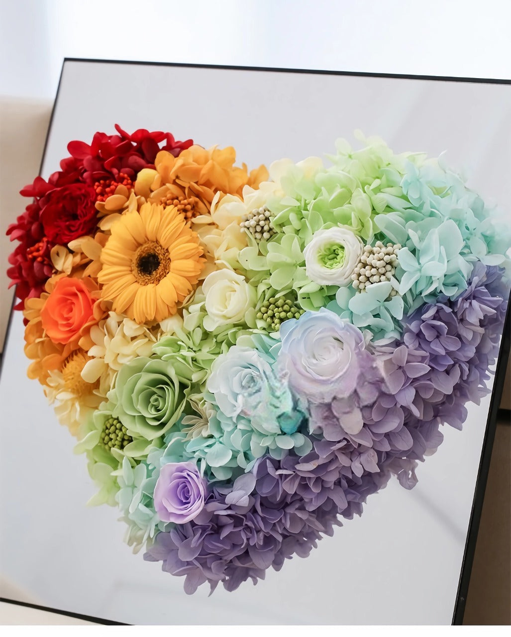 The Rainbow Heart Floral Box features vibrant preserved roses, a sunflower, and hydrangeas arranged in a heart shape. This charming eternity rose gift is perfect for special occasions like Valentine's Day or anniversaries.